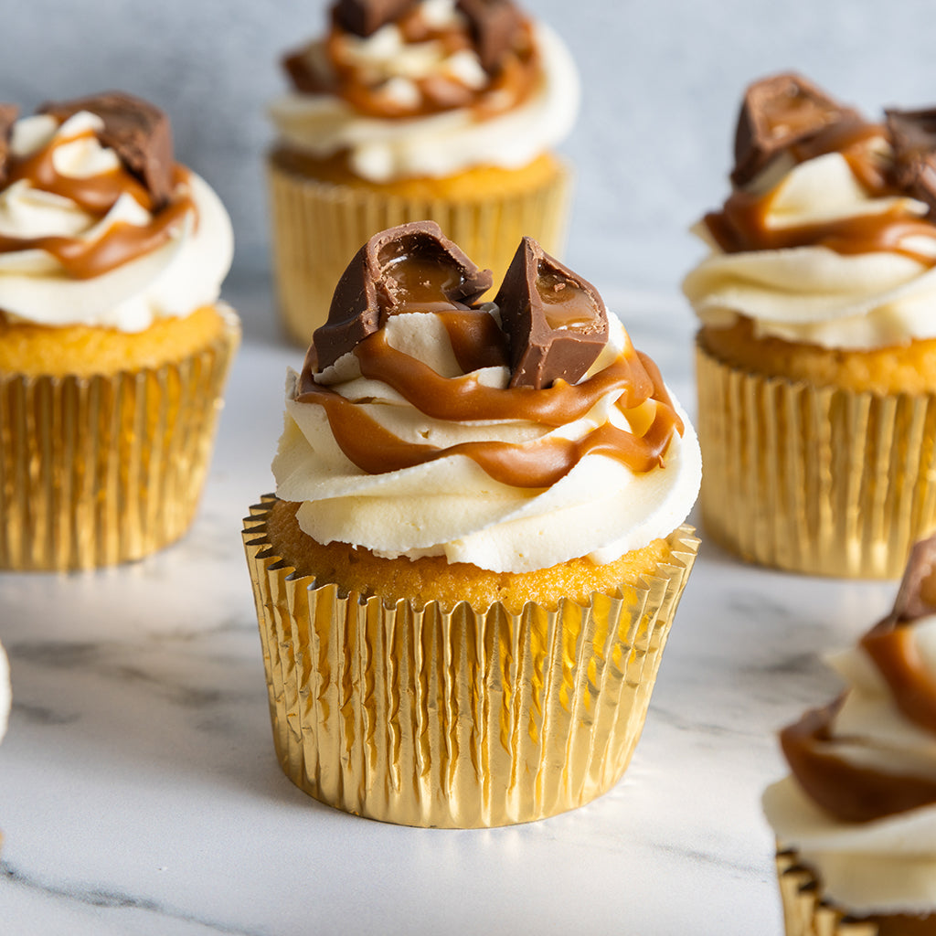 Cupcakes with white frosting, caramel, and chocolate pieces on a marble surface.