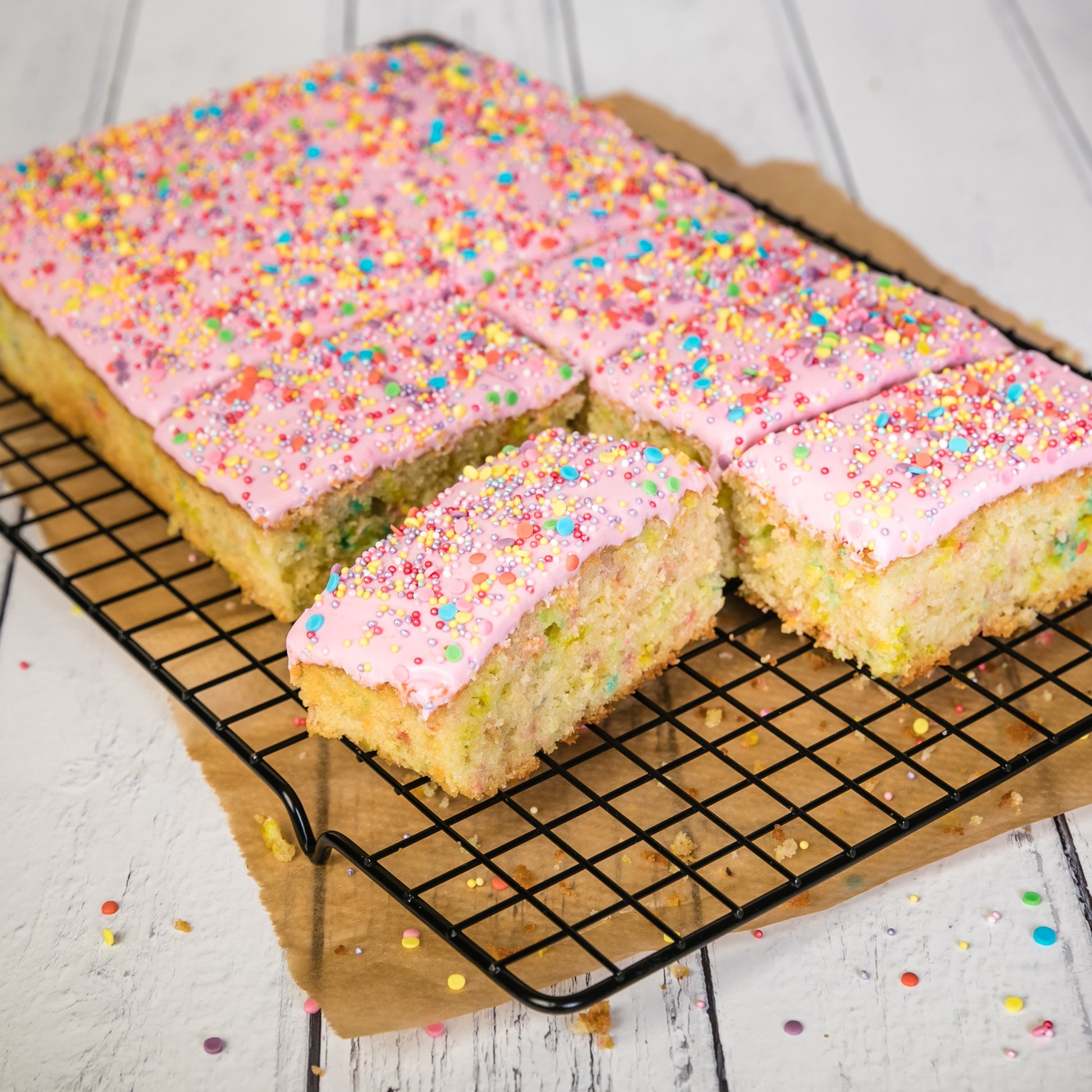 Square slices of cake with pink frosting and colorful sprinkles on a cooling rack.
