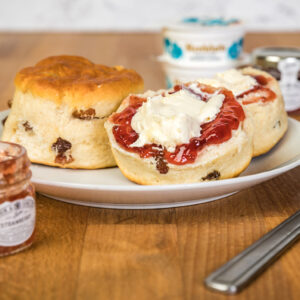 The image shows a classic cream tea setup featuring two fruit scones placed on a white plate. One scone is whole, while the other is halved and generously topped with rich red strawberry jam and a dollop of clotted cream. You can see plump sultanas baked into the golden-brown scones. In the background, there are small jars of Rodda's clotted cream and strawberry jam, reinforcing the traditional British tea-time feel. The plate sits on a warm wooden surface with a butter knife placed beside it, completing the inviting and indulgent presentation.