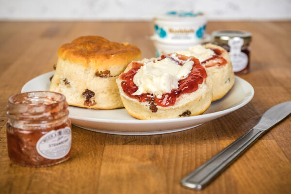 The image shows a classic cream tea setup featuring two fruit scones placed on a white plate. One scone is whole, while the other is halved and generously topped with rich red strawberry jam and a dollop of clotted cream. You can see plump sultanas baked into the golden-brown scones. In the background, there are small jars of Rodda's clotted cream and strawberry jam, reinforcing the traditional British tea-time feel. The plate sits on a warm wooden surface with a butter knife placed beside it, completing the inviting and indulgent presentation.
