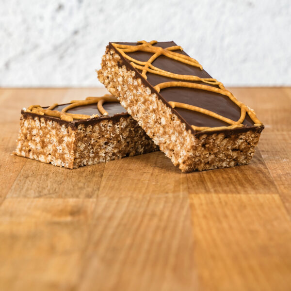 Two Mars Bar slices on a wooden surface, featuring crispy cereal bars topped with a smooth chocolate layer and drizzled with caramel decoration.