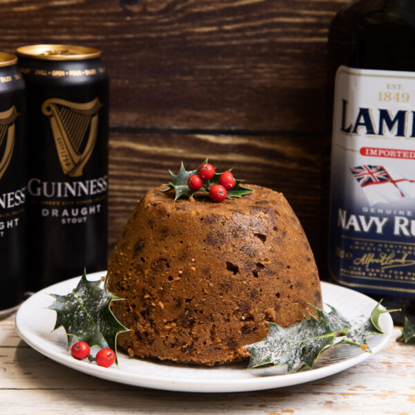 The Warings Christmas Pudding displayed on a plate topped with Holly & berries with bottles of rum and cans of stout in background.