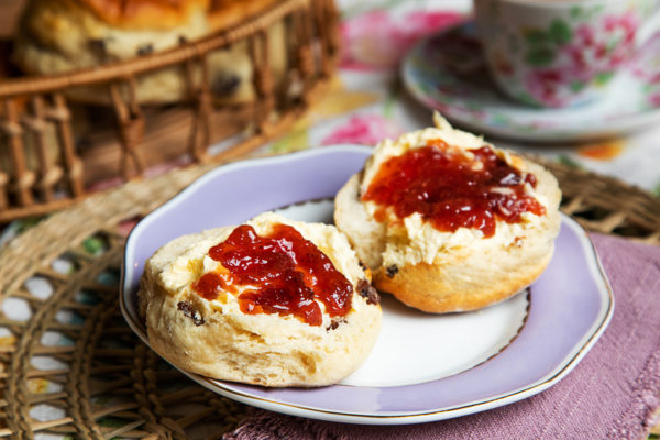 A scone split in two spread with clotted cream and a dollop of strawberry jam on top both halves. The scones are served on a white plate with lilac border and placed on a wicker table mat with lilac cotton napkin