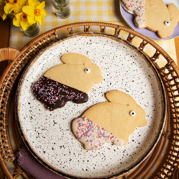 twobunny shaped shortbread biscuits laid flat and displayed on a specled grey plate. One bunny half dipped in white chocolate and sprinkles, the other half dipped in chcocolate and sprinkles