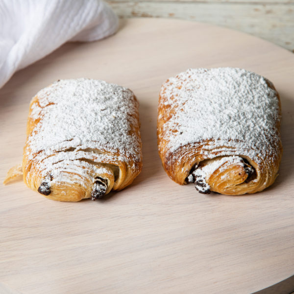 two all butter pain au chocolat pastry dusted in icing sugar display on a light coloured board with tea towel in background