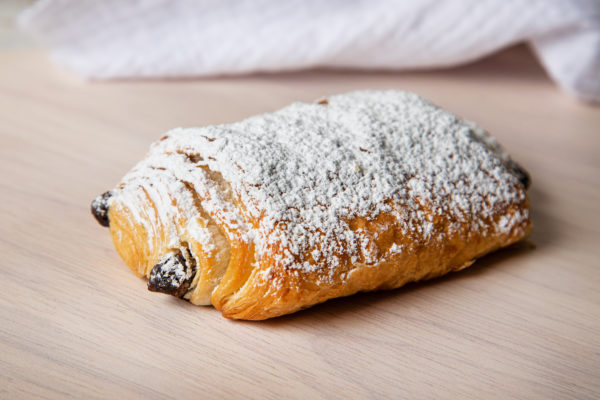 single all butter pain au chocolat pastry dusted in icing sugar display on a light coloured board with tea towel in background