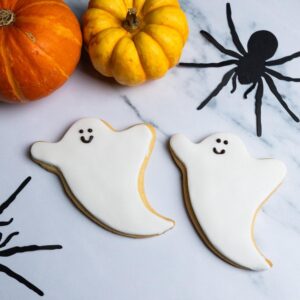 Two ghost-shaped shortbread biscuits covered in white sugar paste with smiling faces, surrounded by small pumpkins and paper spiders on a marble background.