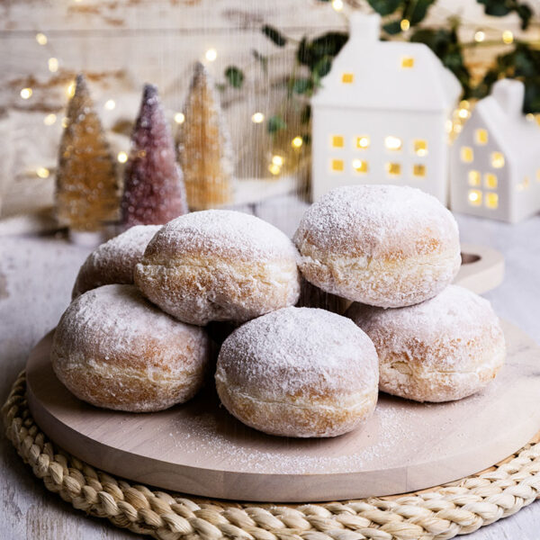 a display of custard snowball doughnuts displayed on a wodding choppingboard with christmas scene in the background of some gingerbread and wooden cut out frosted house