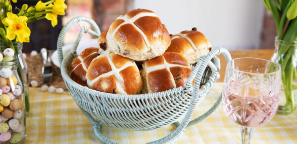 Six Hot Cross Buns displayed in a duck egg blue wicker basket on a gingham table table cloth with daffodils in a vase in the background