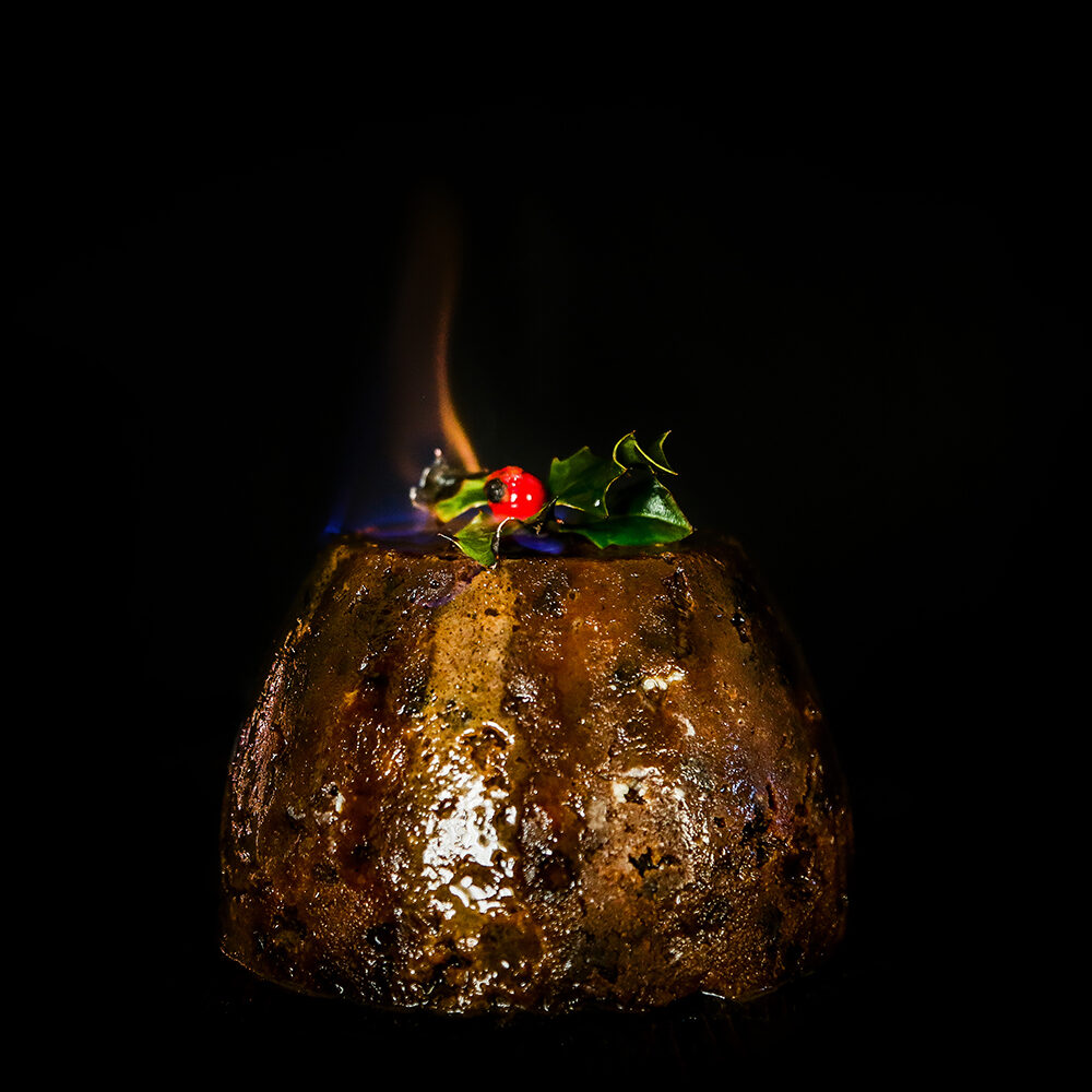 The Warings Christmas Pudding set alight with a small orange and blue flame showing against a black background