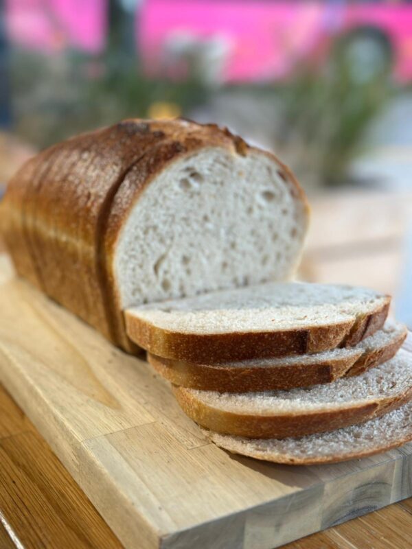 A sliced sourdough farmhouse loaf resting on a wooden chopping board. The bread has a golden-brown crust and a soft, airy interior, with several slices fanned out in the foreground. The background is softly blurred, suggesting a warm, natural light setting.