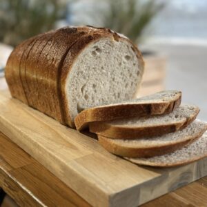 A sliced sourdough farmhouse loaf resting on a wooden chopping board. The bread has a golden-brown crust and a soft, airy interior, with several slices fanned out in the foreground. The background is softly blurred, suggesting a warm, natural light setting.