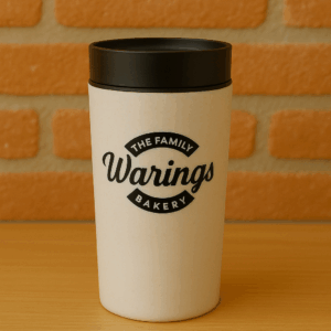 A reusable Circular & Co Warings Bakery travel mug is displayed on a light wooden surface with a rustic red brick wall as the backdrop. The mug has a smooth, off-white speckled finish with a matte black lid and features the bold black Warings logo that reads “The Family Warings Bakery” prominently across the front. The warm, natural tones and clear lighting give the image a cozy and artisanal feel.