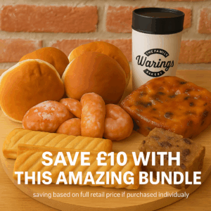 A wooden board displays a selection of Warings Bakery treats, including six soft white round rolls, sugar-glazed double knot doughnuts, two golden flaky sausage roll, a slice of rich bread pudding, and a sticky lardy cake. Behind the baked goods sits a cream reusable travel mug with a black lid, clearly branded with “The Family Warings Bakery” logo. The background features a rustic red brick wall, adding warmth and charm to the scene.
