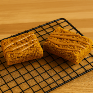 Two golden Biscoff flapjack bars resting on a black wire cooling rack. Each bar is topped with a crumbly Biscoff biscuit layer and finished with a neat drizzle of Biscoff spread. The background features a light wooden surface, creating a warm and inviting setting.
