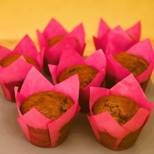 Image showing a group of freshly baked banana and chocolate muffins in vibrant pink tulip-style paper cases. The muffins have a golden-brown top with visible hints of chocolate flakes and are arranged closely together on a neutral surface with a warm yellow background.