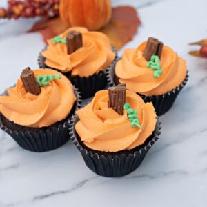 Four chocolate cupcakes decorated with orange buttercream swirls, chocolate flake stems and green icing leaves to resemble pumpkins, displayed on a marble surface with autumn leaves and a small pumpkin decoration.
