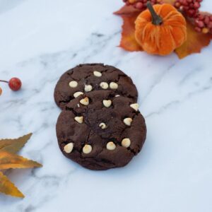 Two double chocolate cookies with white chocolate chips on top, displayed on a marble surface with autumn leaves and a small pumpkin decoration