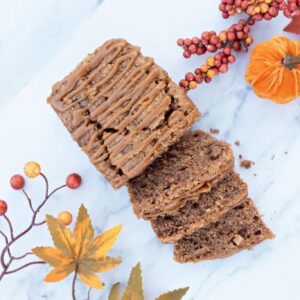 Sticky Toffee Apple Loaf Cake topped with a drizzle of toffee icing, shown sliced on a marble surface with autumn leaves, berries and a small pumpkin decoration.